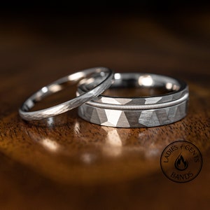 May include: Two silver wedding rings on a wooden surface. One ring has a hammered texture, the other features a geometric design with a guitar string inlay. The rings are likely for a couple.