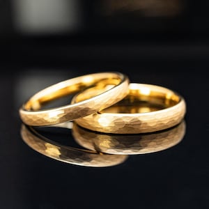 May include: Two gold wedding bands with a hammered texture are displayed on a reflective surface. The rings are a warm gold color, and the texture creates a unique, eye-catching design. The rings are likely for a wedding or engagement.