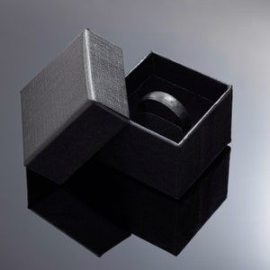 Op de afbeelding: Een zwarte ring in een zwarte, getextureerde geschenkdoos. De ring heeft een gehamerde textuur. De doos is open, waardoor de ring tegen een donkere achtergrond zichtbaar is. De ring is een bandstijl.