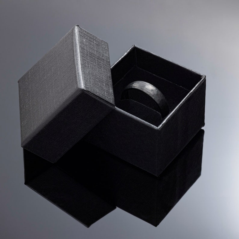 Op de afbeelding: Een zwarte ring in een zwarte, getextureerde geschenkdoos. De ring heeft een gehamerde textuur. De doos is open, waardoor de ring tegen een donkere achtergrond zichtbaar is. De ring is een bandstijl.