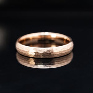 Könnte beinhalten: Ein roségoldener Ring mit gehämmertem Finish. Der Ring hat eine strukturierte Oberfläche mit leicht gewölbter Form. Der Ring wird auf einer schwarzen Oberfläche präsentiert, die seine Form widerspiegelt.