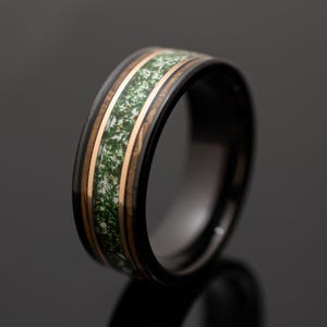 May include: A black band ring featuring a central inlay of white material with green moss-like inclusions, flanked by two thin copper-colored metallic bands that separate the inlay from the polished black outer edges of the ring.