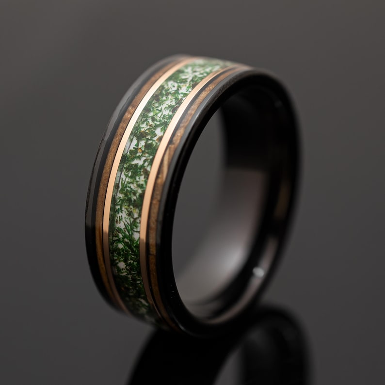 May include: A black band ring featuring a central inlay of white material with green moss-like inclusions, flanked by two thin copper-colored metallic bands that separate the inlay from the polished black outer edges of the ring.