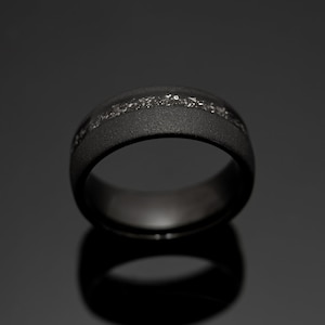 May include: A black ring with a textured surface and a silver-colored inlay. The ring is set against a dark background, highlighting its matte finish and the reflective quality of the inlay.