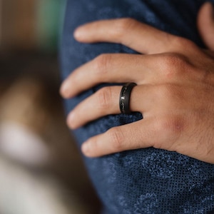 May include: A black ring with a textured surface and a decorative inlay. The ring is worn on a hand, with a blue patterned fabric in the background.