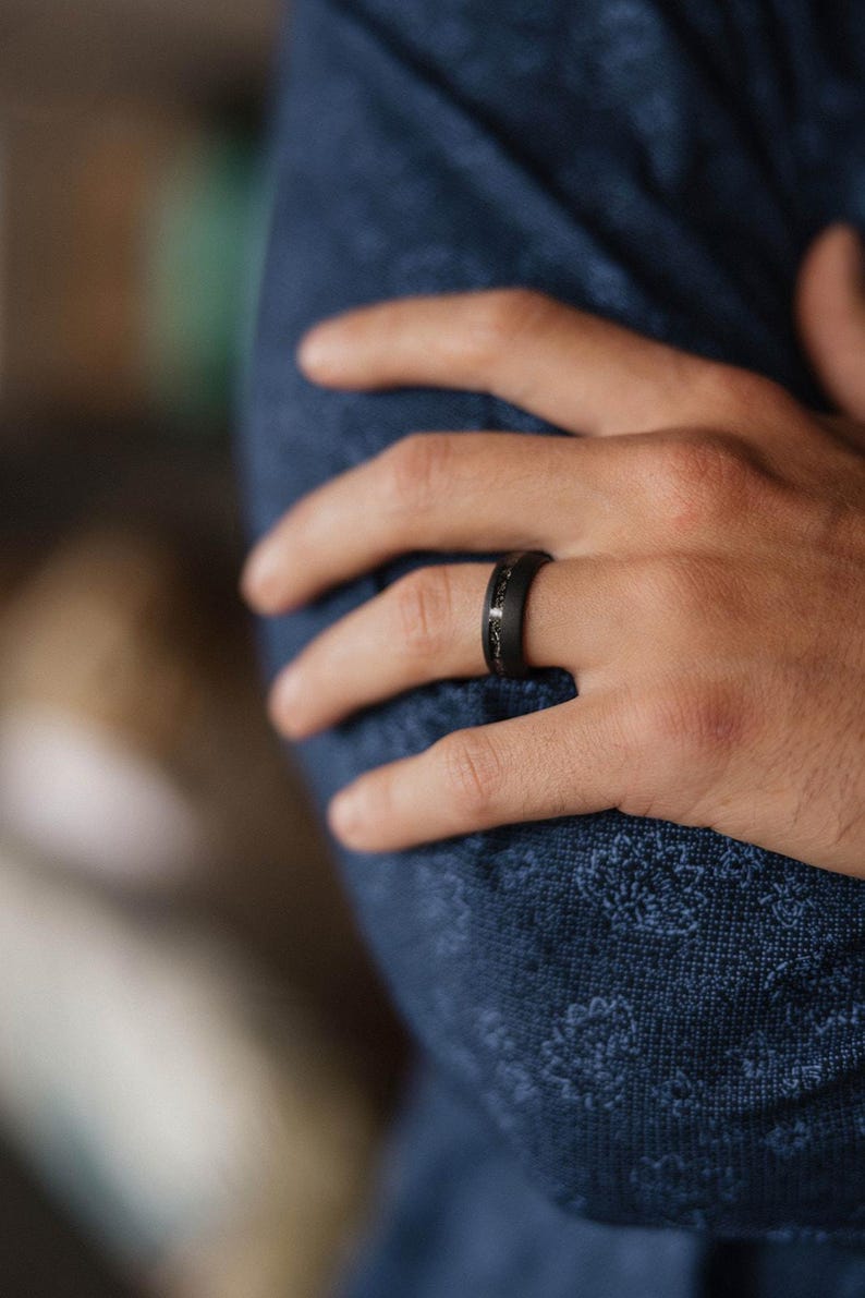 May include: A black ring with a textured surface and a decorative inlay. The ring is worn on a hand, with a blue patterned fabric in the background.