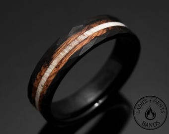 Op de afbeelding: Zwarte ring met een facetgeslepen oppervlak, met een centrale inleg van lichtbruin hout en een witte streep. Het ontwerp van de ring heeft een donkere, getextureerde buitenkant, wat een opvallend contrast creëert met de lichtere binnenelementen. Het is een sieraad.
