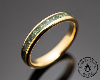 May include: A gold-toned ring featuring a central inlay of shimmering green crushed material. The band has a polished, smooth finish. A circular logo in the bottom right corner contains the text LADIES & GENTS BANDS with a flame icon.