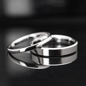 Può includere: Due anelli d'argento, uno sovrapposto all'altro, che si riflettono su una superficie scura. L'anello superiore è sottile e arrotondato, mentre l'anello inferiore è più largo con una superficie piatta. Gli anelli sono molto lucidi e riflettenti.