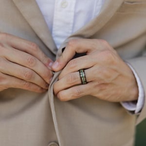 May include: A dark-toned metal ring featuring a central inlay of crushed green and gold-colored stone fragments. The band has a smooth, polished finish and is worn on a finger, positioned against a light beige blazer and white shirt.