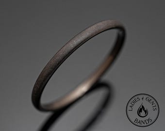 Op de afbeelding: Een dunne, donkergrijze ring met een matte, getextureerde afwerking. De band heeft een eenvoudig, afgerond profiel. In de rechterbenedenhoek staat een cirkelvormig logo met de tekst "LADIES & GENTS BANDS" en een icoon van een vlam.