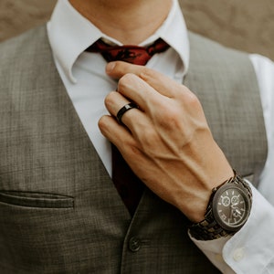 May include: A grey vest worn over a white dress shirt with a dark red tie. A black ring is worn on the finger, and a dark metal chronograph watch with a textured link band is visible on the wrist, completing the formal attire ensemble.