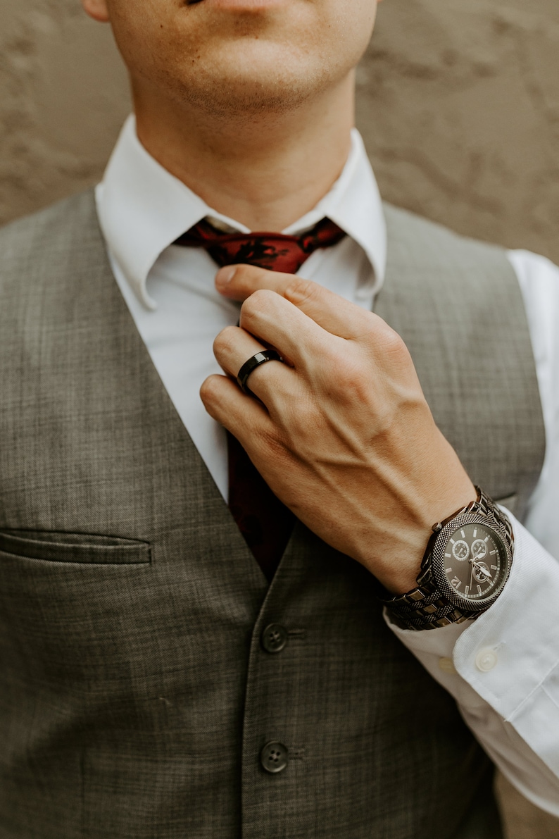 May include: A grey vest worn over a white dress shirt with a dark red tie. A black ring is worn on the finger, and a dark metal chronograph watch with a textured link band is visible on the wrist, completing the formal attire ensemble.