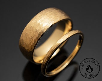 May include: Two gold wedding bands with a hammered texture. The larger band has a wider profile, while the smaller band is thinner. The rings are displayed on a reflective black surface. The image includes a logo that says "Ladies & Gents Bands".