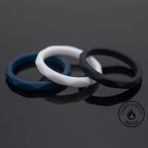 May include: Three silicone rings in navy blue, white, and black, each with a faceted design. The rings are arranged on a reflective black surface. The rings are from Ladies & Gents Bands.