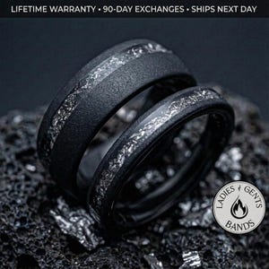 May include: Two black rings with a silver inlay are displayed on a dark, textured surface. The rings have a matte finish and a band of silver-colored material running through the center. The text "LADIES & GENTS BANDS" is visible in a circular logo.
