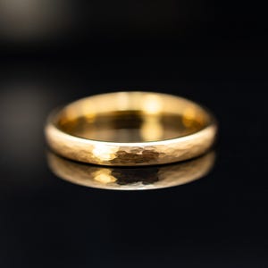 Könnte beinhalten: Nahaufnahme eines goldenen Rings mit gehämmertem Muster. Der Ring liegt vor einem schwarzen Hintergrund, mit seiner Reflexion darunter. Die Oberfläche des Rings weist ein strukturiertes, unregelmäßiges Muster auf.