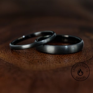 Può includere: Due fedi nuziali grigio scuro, una sottile e una più larga, sono appoggiate su una superficie di legno. Gli anelli hanno una finitura in metallo spazzolato. L'immagine include un logo circolare con la scritta "Ladies & Gents Bands".