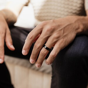 May include: A smooth, polished black band ring with a flat profile and a reflective surface. The ring is worn on the ring finger, showcasing a solid, dark metallic finish that contrasts with the skin tone, creating a clean and simple look.