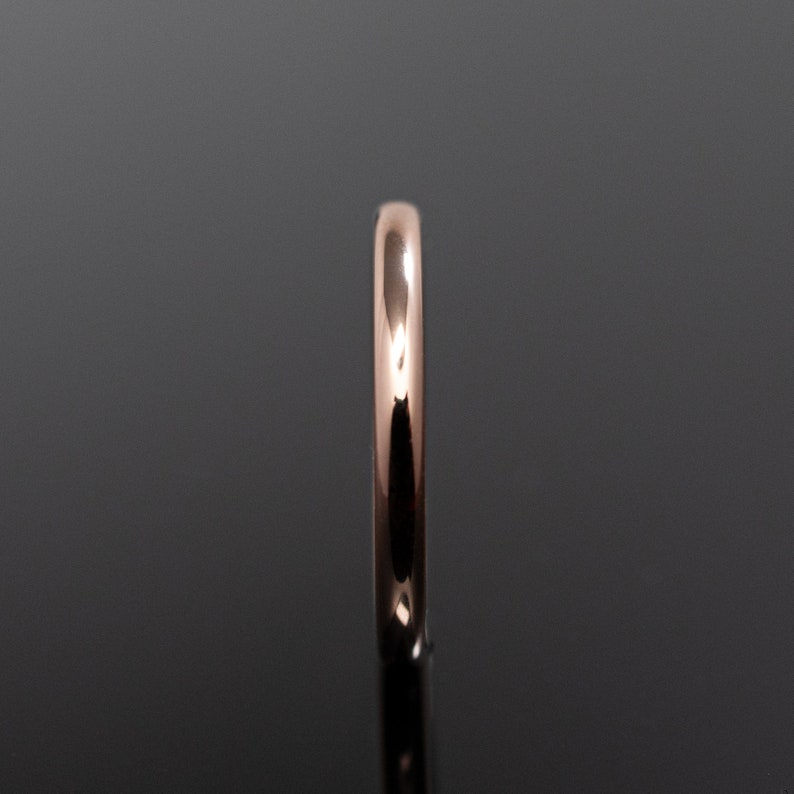 May include: A polished rose gold ring with a smooth, rounded band profile. The metal surface is highly reflective, showing a warm metallic hue and a seamless, continuous circular shape captured from a side-on perspective against a dark background.