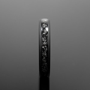 Peut inclure: Gros plan d'une bague noire avec une bande incrust&eacute;e de fragments scintillants de forme irr&eacute;guli&egrave;re. La bague est pos&eacute;e sur un fond sombre et r&eacute;fl&eacute;chissant, soulignant le contraste entre la bande mate et les fragments brillants.