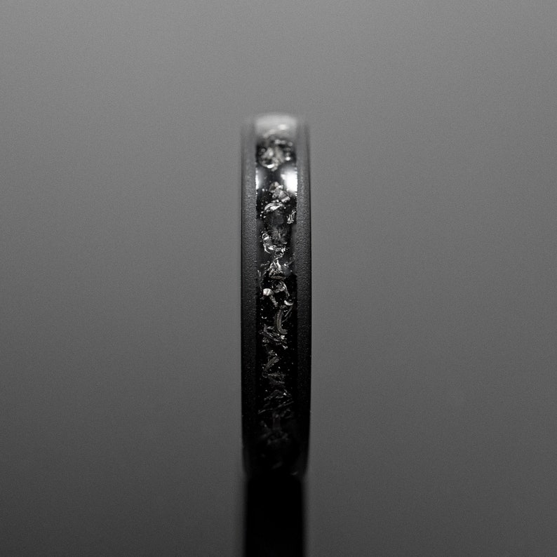 Peut inclure: Gros plan d'une bague noire avec une bande incrust&eacute;e de fragments scintillants de forme irr&eacute;guli&egrave;re. La bague est pos&eacute;e sur un fond sombre et r&eacute;fl&eacute;chissant, soulignant le contraste entre la bande mate et les fragments brillants.