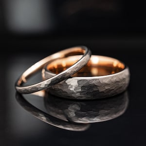 Puede incluir: Dos anillos de boda. Uno es una banda delgada plateada, el otro es una banda más ancha con una textura martillada. El interior del anillo más ancho es dorado. Los anillos están sobre una superficie negra reflectante.