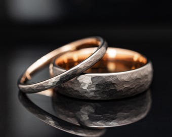 May include: Two rings with a hammered exterior texture and a smooth rose gold interior. The wider band is positioned behind the thinner band, both featuring a dark grey metallic finish on the outer surface and a polished rose gold lining.
