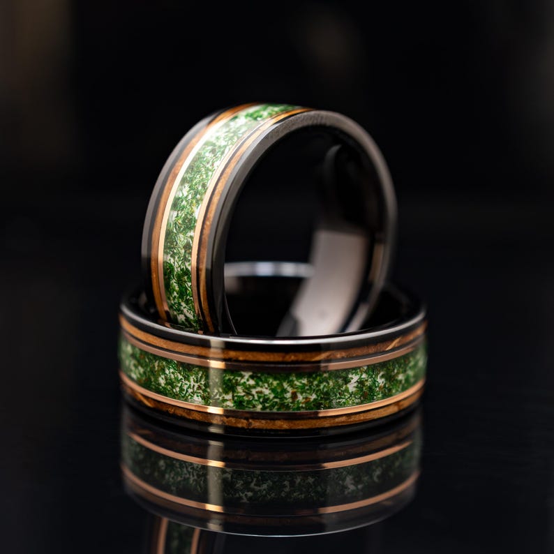 May include: A black ring featuring a central inlay of green moss-like material flanked by two thin copper-colored bands. The ring has a smooth, polished finish and is positioned upright, with a second identical ring lying flat beneath it.