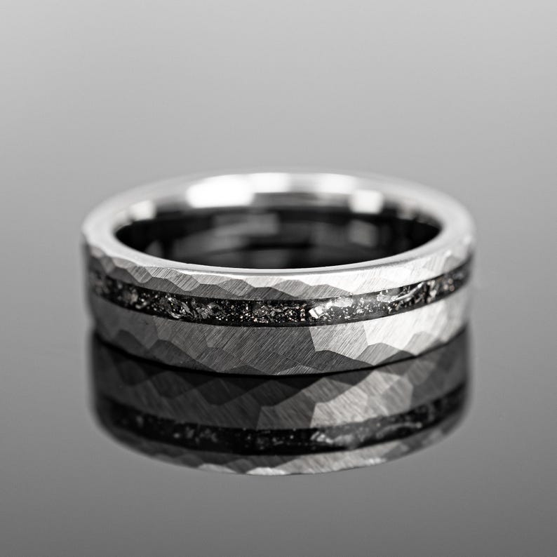 May include: A silver ring with a textured, hammered finish and a dark, speckled inlay. The ring features a wide band and a polished surface, reflecting light. The design is modern and minimalist, suitable for everyday wear or special occasions.