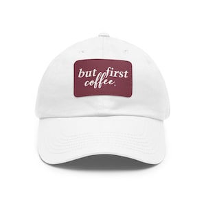 Op de afbeelding: Witte baseballpet met een bordeauxrode patch waarop "but first coffee." staat.