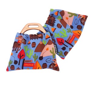May include: Two small blue drawstring bags with a wooden handle. The bags feature a repeating pattern of cowboys, cacti, mountains, and the words "GUNSHOP SALOON". The bags are designed with a western theme.