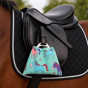 May include: A teal drawstring bag with a unicorn pattern hangs from a black leather saddle on a horse. The bag features pink, blue, and purple unicorns with polka dots and red manes. The saddle pad is black with a white border.