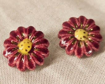 May include: A pair of ceramic flower earrings with a burgundy red base and a yellow center. The flowers have gold accents around the edges.