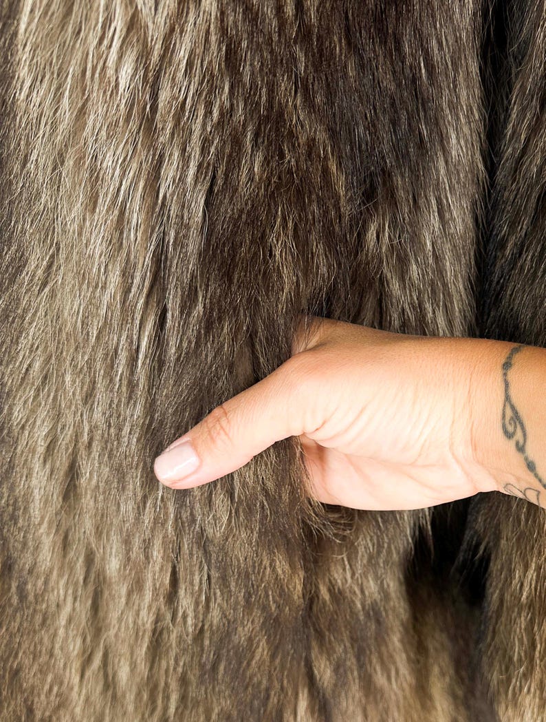 May include: Close-up of a brown and gray fur textile. The fur has a dense, textured appearance with long, flowing strands. A hand with a tattoo is touching the fur, highlighting its softness and texture.