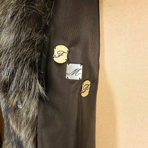 May include: Close-up of a dark brown coat interior with a fur collar. The coat features three small decorative tags with the letters "F", "M", and "P". The tags are in gold and silver tones.