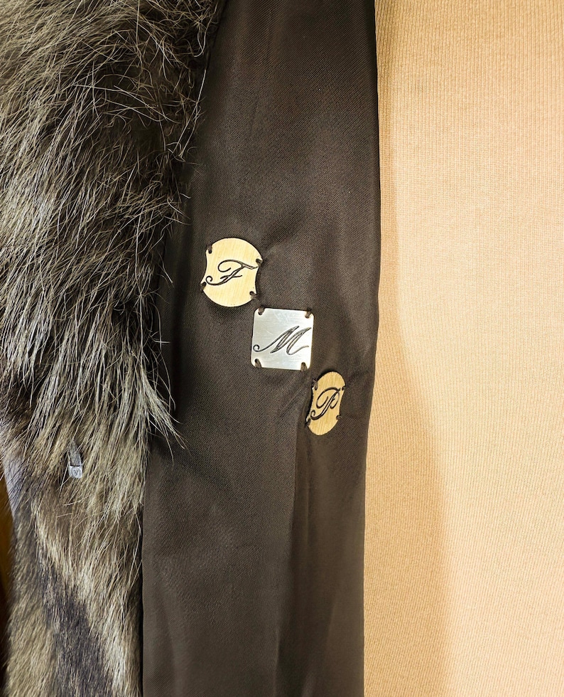 May include: Close-up of a dark brown coat interior with a fur collar. The coat features three small decorative tags with the letters "F", "M", and "P". The tags are in gold and silver tones.