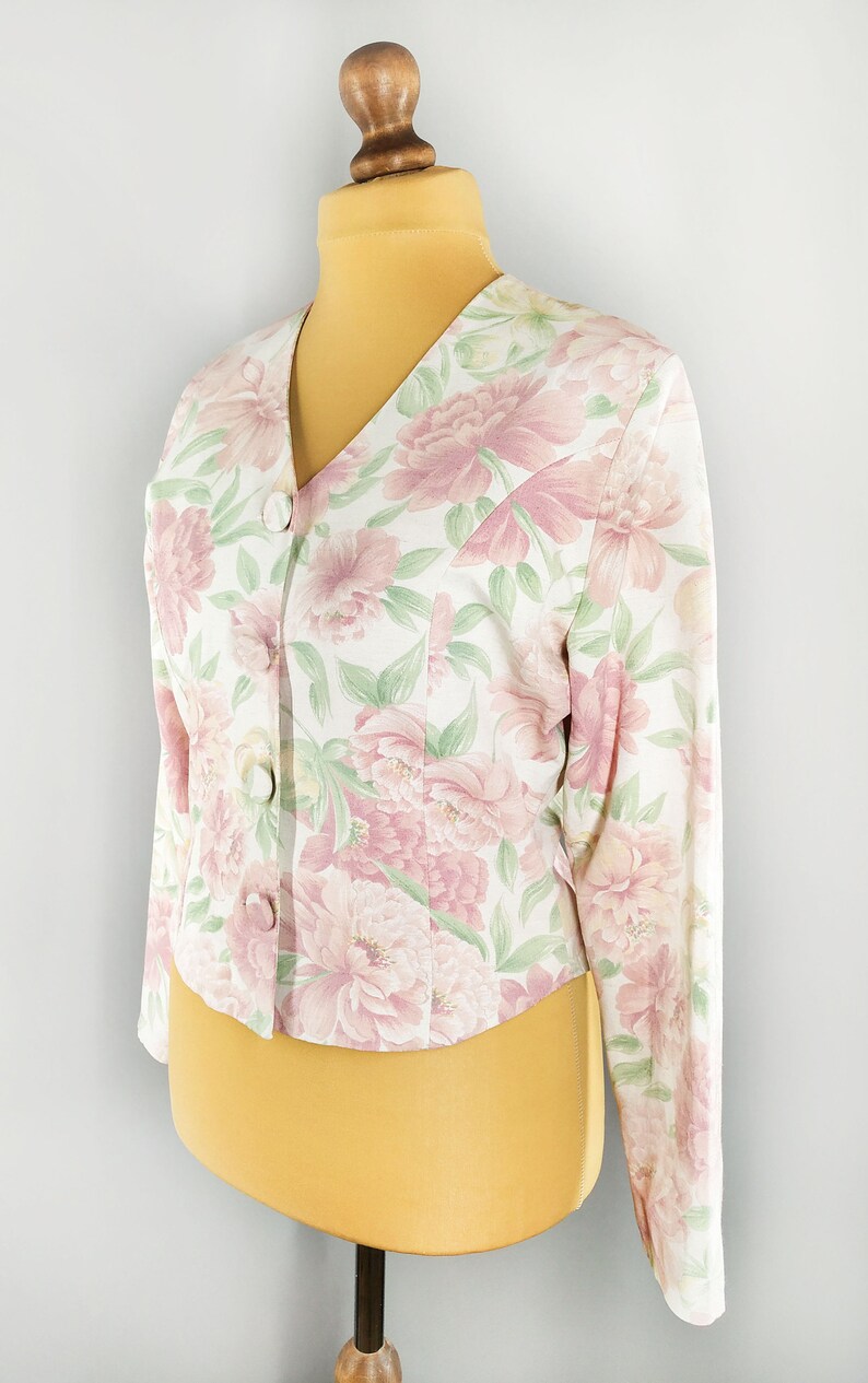 May include: A white floral print blazer with pink flowers and green leaves. The blazer has a v-neck and long sleeves.
