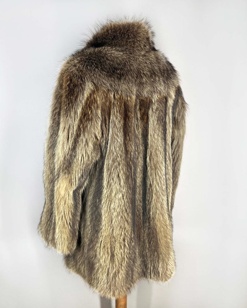 May include: A vintage fur coat with a natural color palette of browns and tans. The coat has a wide collar and a long, flowing design. The fur appears to be in good condition, with a soft and textured appearance. The coat is displayed on a wooden stand.