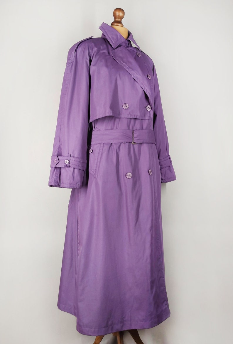 May include: A long, purple trench coat with a double-breasted closure, a belt, and a collar. The coat has a classic design and is made of a smooth, lightweight fabric.