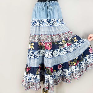 May include: A long, tiered skirt with a denim waistband and drawstring. The skirt features alternating panels of floral, gingham, and patterned fabrics in shades of blue, white, and pink. The skirt has a flowing, bohemian style.