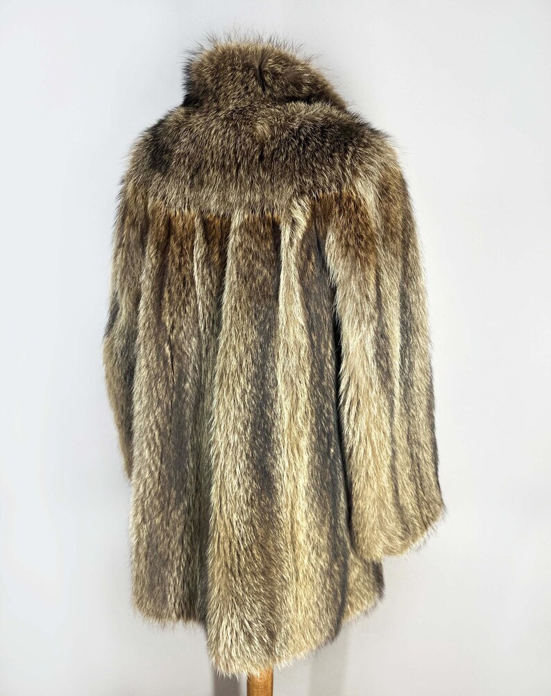 May include: A vintage fur coat in shades of brown and tan, with a textured, striped pattern. The coat has a collar and long sleeves, and appears to be in good condition. The coat is displayed on a wooden hanger against a white background.