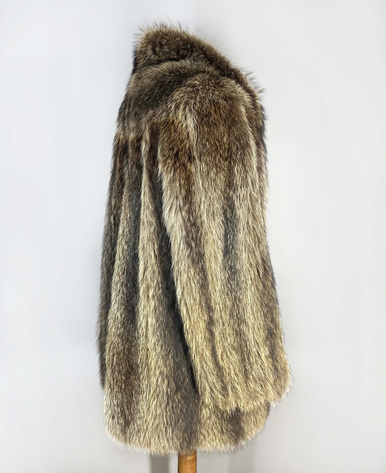 May include: A long, faux fur coat in shades of brown and tan. The coat has a collar and long sleeves. The fur appears soft and plush, with a natural-looking texture. The coat is displayed on a wooden hanger.