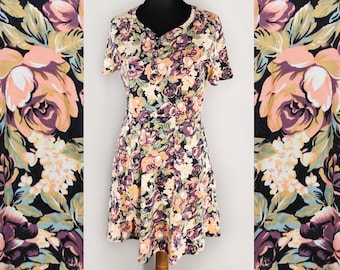 Puede incluir: Un vestido de manga corta con estampado floral en tonos rosa, morado, verde y amarillo sobre fondo negro. El vestido tiene un escote redondo y un cierre de botón en la parte delantera.