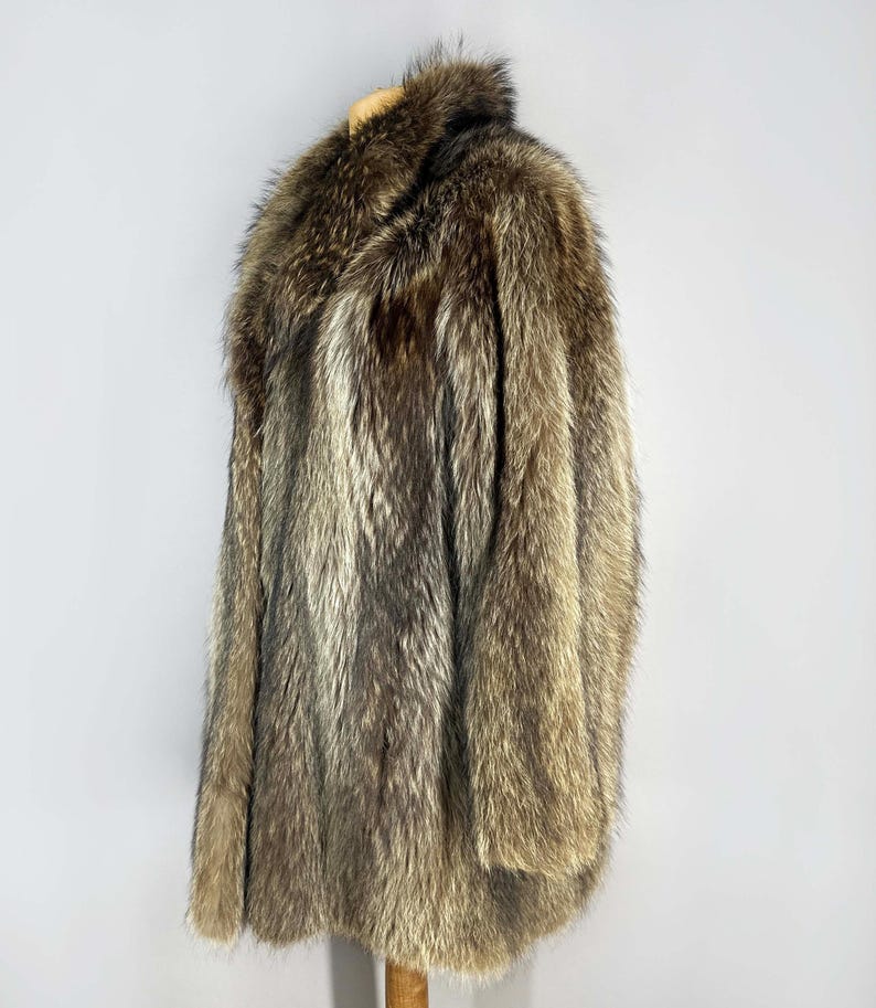 May include: A full-length fur coat in shades of brown, gray, and tan. The coat has a wide collar and long sleeves. The fur appears thick and luxurious, with a natural, textured appearance. The coat is displayed on a wooden hanger.