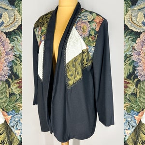 Könnte beinhalten: Eine schwarze Jacke mit offenem Vorderteil und einem floralen Wandteppichmuster auf der Vorderseite. Der Wandteppich besteht aus verschiedenen Stoffen in Farben wie Grün, Weiß und Braun.