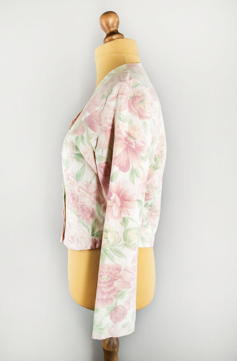 May include: A white cropped jacket with a pink and green floral print. The jacket has long sleeves and a button-up front.