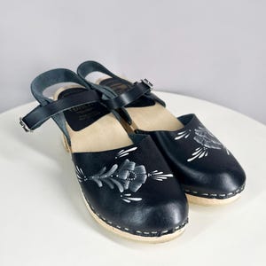 May include: A pair of black leather clogs with white floral designs. The clogs have a wooden base and an ankle strap with a buckle.