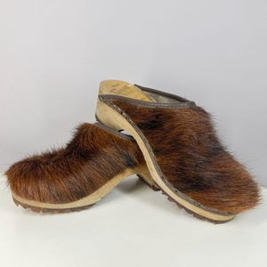 Könnte beinhalten: Ein Paar braune Fellclogs mit Holzsohlen. Die Clogs haben ein Obermaterial aus dunkelbraunem Fell und eine hellbraune Holzbasis. Die Sohlen haben ein geriffeltes Design für Traktion. Die Clogs sind leicht angewinkelt.
