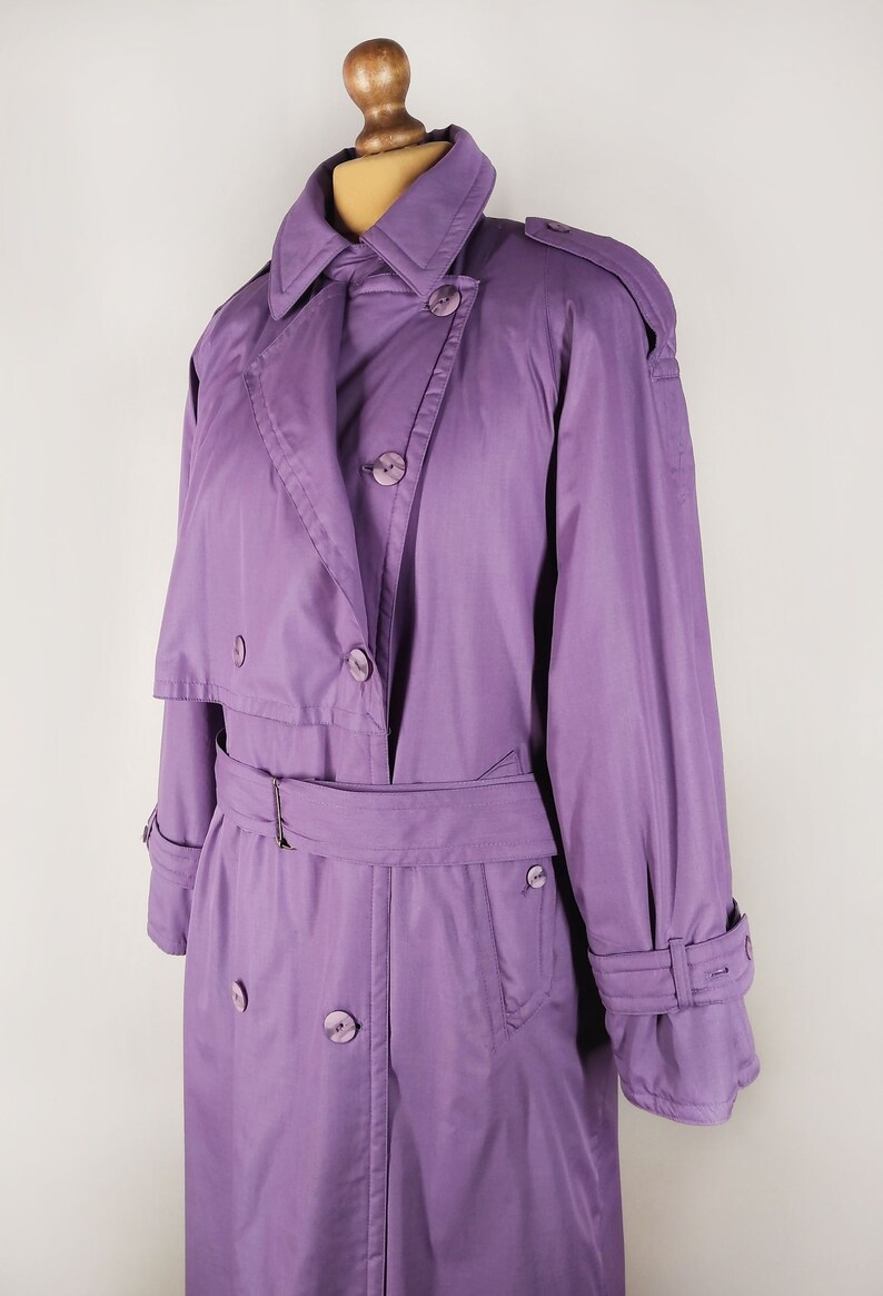 May include: A long, purple raincoat with a double-breasted closure, a belt, and a storm flap. The raincoat has a classic trench coat style.